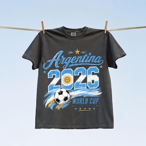 May include: A charcoal grey short-sleeve t-shirt featuring a graphic print with the text Argentina 2026 World Cup. The design includes a football, the Argentine flag colours, a sun emblem, and five small stars in a light blue and white colour palette.