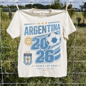 May include: A cream-coloured t-shirt featuring a blue graphic print that reads ARGENTINA 2026, WORLD CUP 2026, and ROAD TO THE CUP. The design includes a football, a sun emblem, and the Argentina national team crest with two gold stars above it.