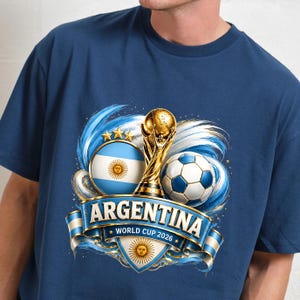 May include: A navy blue t-shirt featuring a graphic of the Argentina flag, a gold trophy, and a football. A blue banner across the centre displays the text ARGENTINA and WORLD CUP 2026, with a sun emblem below the text and three gold stars above.