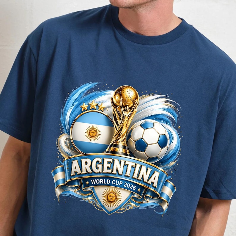 May include: A navy blue t-shirt featuring a graphic of the Argentina flag, a gold trophy, and a football. A blue banner across the centre displays the text ARGENTINA and WORLD CUP 2026, with a sun emblem below the text and three gold stars above.