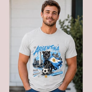 May include: A light grey t-shirt featuring a graphic of a black panther draped in an Argentine flag, a football, the Obelisco de Buenos Aires, and the text "ARGENTINA", "PASI&Oacute;N ORGULLO ALBICELESTE", and "VAMOS ARGENTINA" in blue and black ink.
