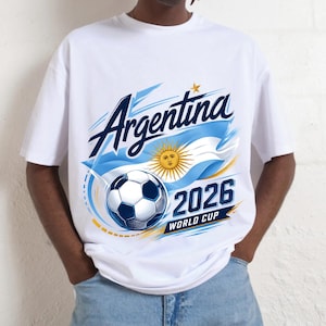 May include: A white t-shirt featuring a graphic print with the text Argentina, 2026, and World Cup. The design includes a football, a stylised sun, and a blue and white flag motif with yellow accents, centred on the front of the garment.
