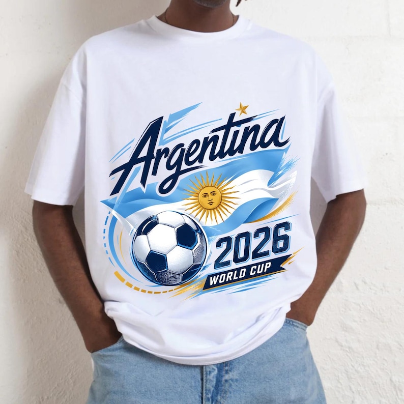 May include: A white t-shirt featuring a graphic print with the text Argentina, 2026, and World Cup. The design includes a football, a stylised sun, and a blue and white flag motif with yellow accents, centred on the front of the garment.