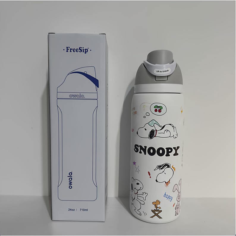 May include: White Owala FreeSip water bottle with a gray lid and Snoopy cartoon graphics. The bottle has a 24oz capacity. A matching white box with the Owala logo and bottle outline is on the left.