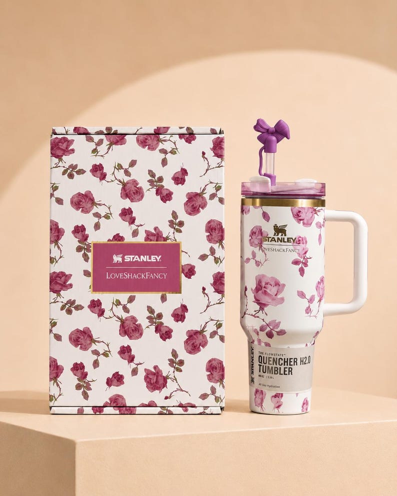May include: A white Stanley Quencher H2.0 tumbler and its matching box, both featuring a pink rose floral pattern. The tumbler has a white handle, a gold rim, and a purple bow-shaped straw topper. The box displays the Stanley and LoveShackFancy logos.