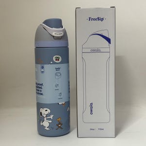 May include: A blue and gray Owala FreeSip water bottle with a Snoopy design, next to its matching box. The bottle has a flip-top lid and a straw. The box reads "FreeSip" and "owala." The bottle holds 24 oz.