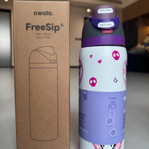Puede incluir: Una botella de agua blanca Owala FreeSip con tapa y detalles morados, con un dise&ntilde;o de calaveras y corazones. La botella est&aacute; junto a su caja de cart&oacute;n, que tiene el nombre de la marca y la ilustraci&oacute;n de la botella.