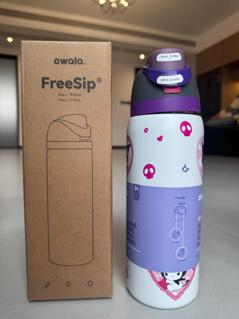 Puede incluir: Una botella de agua blanca Owala FreeSip con tapa y detalles morados, con un dise&ntilde;o de calaveras y corazones. La botella est&aacute; junto a su caja de cart&oacute;n, que tiene el nombre de la marca y la ilustraci&oacute;n de la botella.
