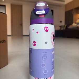 Puede incluir: Una botella de agua blanca y morada con un dise&ntilde;o de dibujos animados. La botella tiene una tapa con bot&oacute;n y una boquilla FreeSip. El dise&ntilde;o incluye calaveras rosas y un personaje de dibujos animados. La botella es de 946 ml.