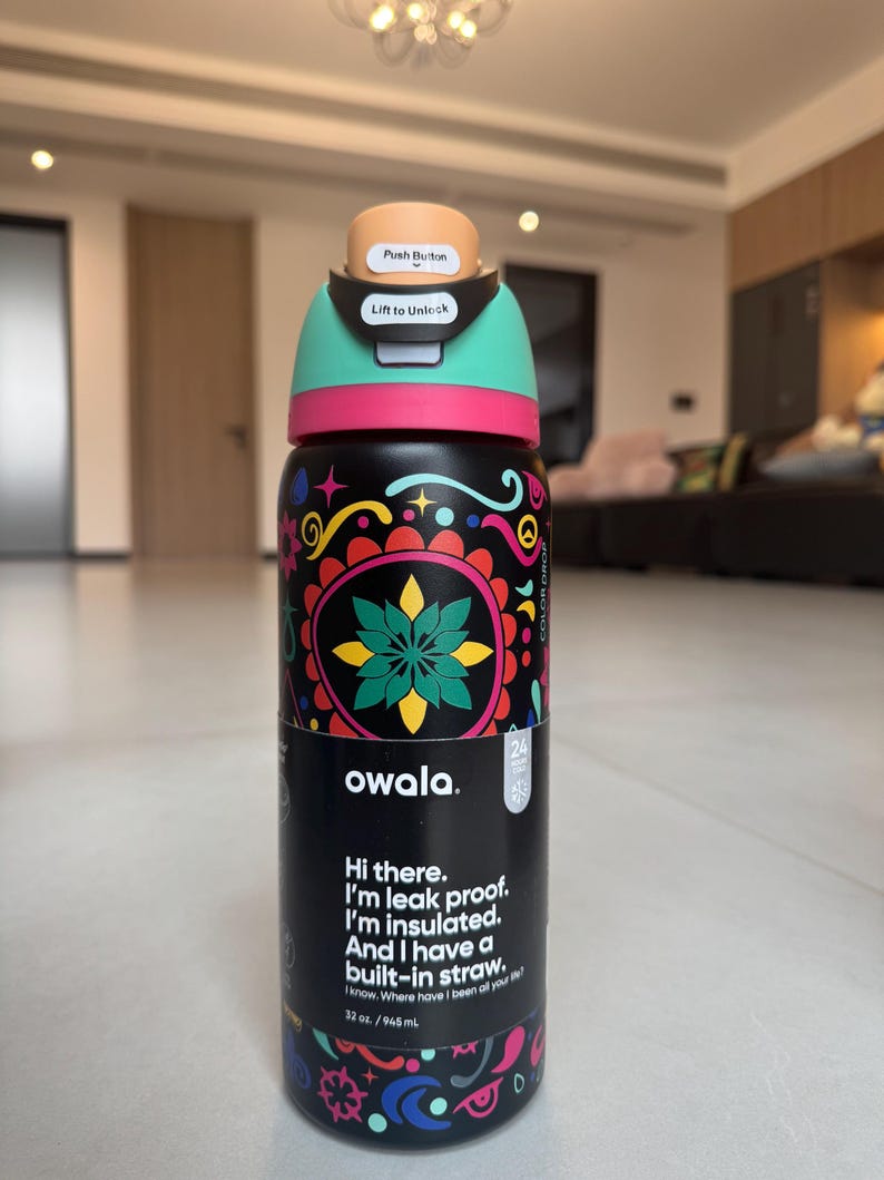 May include: A black Owala water bottle with a vibrant floral pattern. The bottle features a teal lid with a push button and a pink band. Text on the bottle reads, "Hi there. I'm leak proof. I'm insulated. And I have a built-in straw." It holds 945 ml.