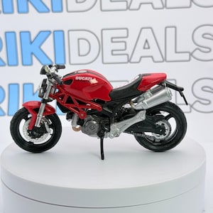 May include: A red Ducati Monster motorcycle model with a black frame, black wheels, and silver exhaust pipes. The fuel tank features the white Ducati logo. The motorcycle is displayed on a white circular stand against a white background with the text RIKIDEALS.