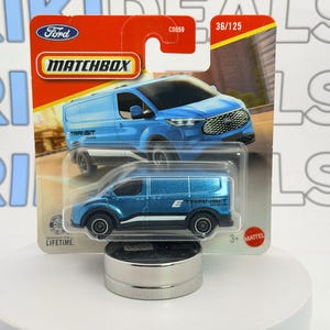 May include: A Matchbox Ford Transit van toy in a blue and silver color scheme. The toy is displayed on a white surface, with the packaging visible. The packaging includes the Ford logo and the Matchbox logo.