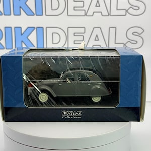 Può includere: Un'auto modello grigia in una scatola blu e nera. L'auto ha un design classico con carrozzeria arrotondata e ruote piccole. La scatola riporta la scritta "EDITIONS ATLAS Collections".