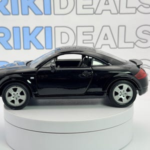Puede incluir: Un coche de juguete negro con ruedas plateadas y un alerón. El coche está sobre un expositor giratorio blanco. El fondo tiene el texto "RIKI DEALS" en azul y gris.