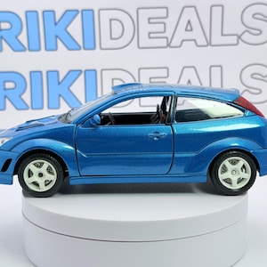 May include: A metallic blue die-cast toy car featuring a three-door hatchback body style, white five-spoke wheels, and a visible black interior. The car is positioned in front of a white background with the repeating text RIKIDEALS in blue and grey.