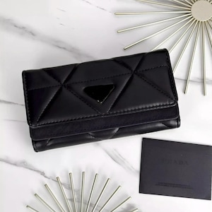 May include: A black quilted wallet with a triangular emblem on the front flap. The wallet features a geometric stitched pattern and a smooth finish. Included is a small black rectangular card with the text PRADA printed in the upper center.