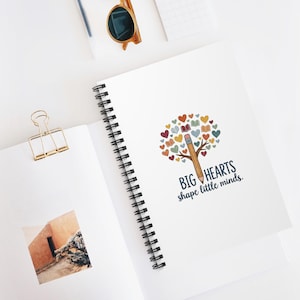 Könnte beinhalten: Weißes Spiralnotizbuch mit einer farbenfrohen Illustration eines Baumes aus Herzen und Büchern sowie einem Bleistiftstamm. Der Text lautet "BIG HEARTS shape little minds." Eine goldene Büroklammer und Sonnenbrille sind ebenfalls sichtbar.