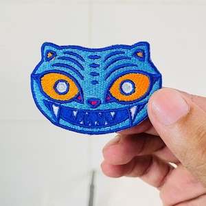May include: An embroidered patch shaped like a stylized cat head in shades of blue. The design features large, bright orange eyes with white pupils, a small pink nose, and a wide mouth displaying sharp white teeth against a dark blue background.