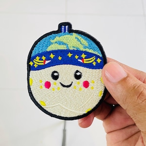 May include: A circular embroidered patch featuring a smiling moon character with a cream-colored face, pink cheeks, and yellow freckles. The moon wears a blue cap decorated with yellow stars, shooting stars, and a light green crescent moon shape.