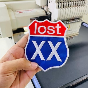 May include: A shield-shaped embroidered patch featuring a red top section with the white text "lost" and a blue bottom section with the large white Roman numerals "XX". The patch has a white stitched border and a clean, finished edge design.