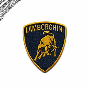 May include: A shield-shaped embroidered patch featuring a dark navy blue background with a golden yellow border. The word LAMBORGHINI is stitched in golden yellow capital letters at the top, above a golden yellow bull silhouette in the center.