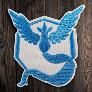 May include: A fabric patch featuring an embroidered design of a stylized blue bird with outstretched wings and a long tail, centered on a white hexagonal background. The embroidery uses bright blue thread to create the detailed avian shape.