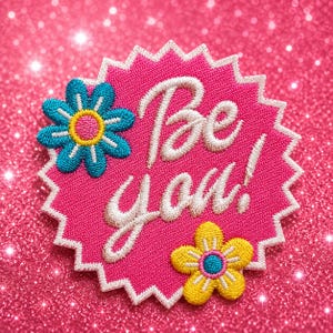 May include: A circular, starburst-shaped embroidered patch with a bright pink background and a white stitched border. The text 'Be you!' is embroidered in white script. A blue flower with a yellow center and a yellow flower with a blue center are attached.