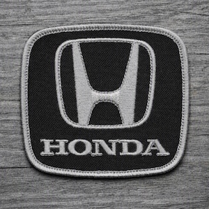 May include: A square-shaped embroidered patch with rounded corners featuring a black background and a silver-colored Honda logo. The iconic H emblem is centered above the word HONDA, both stitched in silver thread with a matching silver border.