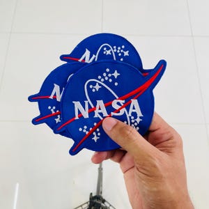 May include: Three circular embroidered patches featuring the blue NASA logo with white lettering, a red swoosh, and white stars on a blue background. The patches are stacked, showing the iconic space agency emblem with detailed stitching.