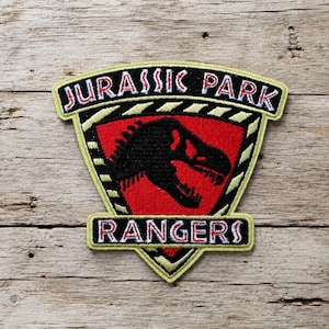 May include: A shield-shaped embroidered patch featuring a black dinosaur silhouette on a red background. The top banner reads JURASSIC PARK in white letters with red outlines, and the bottom banner reads RANGERS in white letters with red outlines.