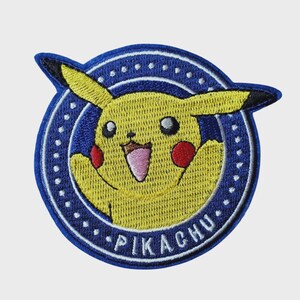 May include: A circular embroidered patch featuring a yellow Pikachu character with red cheeks and black-tipped ears. The character is set against a dark blue background with a white dotted border and the text PIKACHU embroidered in white at the bottom.