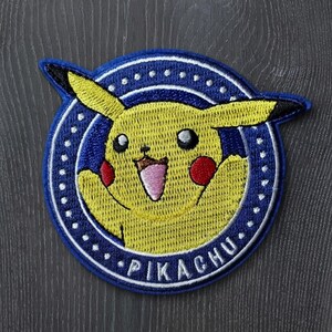 May include: A circular embroidered patch featuring a yellow Pikachu character with red cheeks and a wide open mouth. The character is centered within a dark blue circular border with white dots and the text PIKACHU embroidered in white at the bottom.