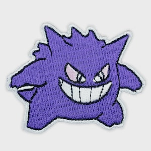 May include: A purple embroidered patch featuring a stylized creature with a wide, toothy grin, pointed ears, and spiky hair. The design includes white eyes with pink accents and a large white mouth with black outlines, all bordered by a white edge.