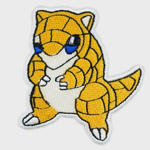 May include: An embroidered patch featuring a stylized yellow creature with a segmented, armor-like body, a white belly, and small white claws. The creature has dark eyes with blue accents and a pointed tail, all outlined in black thread on a white border.