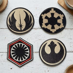 May include: Four embroidered patches featuring science fiction symbols. Three are circular, including a tan and black Jedi symbol, a tan and black Imperial crest, and a tan and black Rebel Alliance logo. One is a black, white, and red hexagonal patch.