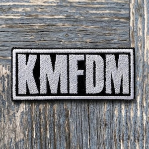 May include: A rectangular embroidered patch featuring the text KMFDM in bold, white, sans-serif capital letters. The lettering is set against a solid black background, and the entire patch is framed by a thick, white, stitched border.