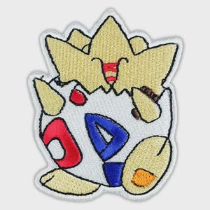 May include: An embroidered patch featuring a cartoon creature with a pale yellow crown-like top and a white egg-shaped body. The body displays red and blue triangular patterns, with small yellow limbs and a small orange oval detail on the lower right side.