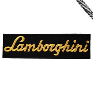 May include: A rectangular black embroidered patch featuring the word Lamborghini stitched in a gold-colored thread in a cursive script font. The patch has a clean, straight-edged border and a flat, fabric-like texture throughout the design.