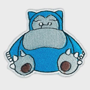May include: An embroidered patch featuring a cartoon creature with a large, round, light blue body, a pale gray belly, and a matching gray face. The character has small ears, closed eyes, and large feet with brown circular pads on the soles.