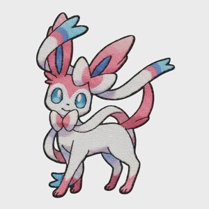 May include: A fabric patch featuring a stylized creature with a white body, pink accents, and large blue eyes. The creature has long, ribbon-like appendages with blue and pink tips, a bow-like neck feature, and tall ears with pink and blue details.