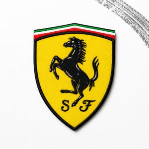 May include: A shield-shaped embroidered patch featuring a yellow background with a black prancing horse silhouette in the center. The letters S and F are stitched in black at the bottom, with a horizontal green, white, and red stripe at the top.