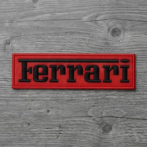 May include: A rectangular embroidered patch featuring a vibrant red background with a thick black border. The word Ferrari is stitched in bold black lettering across the center, with a horizontal black line extending above the text.