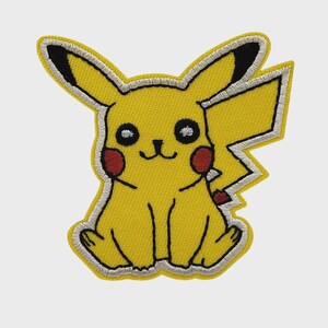 May include: An embroidered patch featuring a yellow creature with long ears tipped in black, red circular cheeks, and a jagged tail. The design includes black outlines, white highlights in the eyes, and a white border surrounding the entire shape.