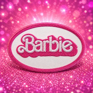 May include: An oval-shaped embroidered patch featuring the white Barbie logo with a thick pink outline. The patch has a white center background and a vibrant pink stitched border, showcasing the iconic script lettering in a bold, raised texture.