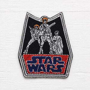 May include: A shield-shaped embroidered patch featuring a black background with white stitched outlines of three figures. Below the figures, a red trapezoid contains the dark blue text STAR WARS. The entire patch has a white stitched border.