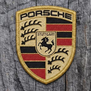 May include: A shield-shaped embroidered patch featuring the Porsche logo in black text at the top. The design includes a central crest with a black horse and the word Stuttgart, flanked by alternating black and red horizontal stripes and gold antlers.