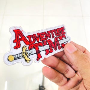 May include: An embroidered fabric patch featuring the text Adventure Time in bold red lettering with a white outline. A silver sword with a yellow hilt is positioned horizontally across the center of the text, integrated into the design.