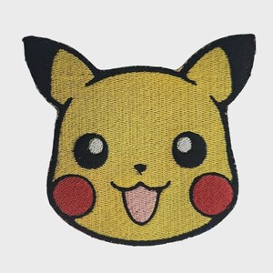 May include: An embroidered patch featuring the head of a yellow creature with pointed ears tipped in black, large black eyes with white highlights, round red cheeks, and a small mouth showing a light pink tongue against a yellow background.