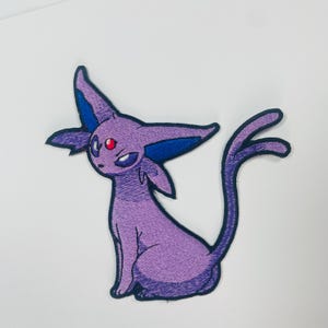 May include: An embroidered patch featuring a stylized purple creature with large pointed ears, dark blue inner ear details, and a small red gem on its forehead. The creature has a slender body, a bifurcated tail, and dark outlines throughout the design.