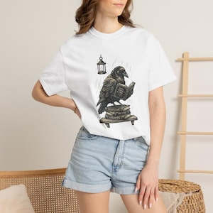 Puede incluir: Una camiseta blanca de manga corta con una ilustración gráfica de un cuervo negro posado sobre una pila de libros. El cuervo sostiene un libro abierto titulado Once Upon A Time, sobre libros etiquetados como Folklore, Myths y Legends.